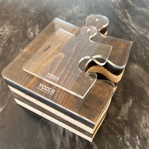 May include: A stack of four wooden puzzle pieces with a clear acrylic puzzle piece on top. The wooden pieces are stained dark brown and have a light wood grain pattern. The acrylic piece has the text "YODER MADE" printed on it.