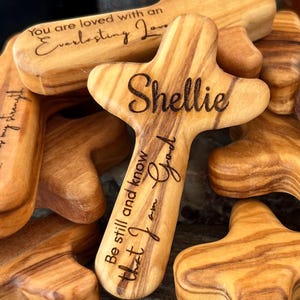 May include: A collection of wooden crosses with rounded edges and a natural wood grain. One cross is engraved with the name "Shellie" and the phrase "Be still and know that I am God." Another reads "You are loved with an Everlasting Love."