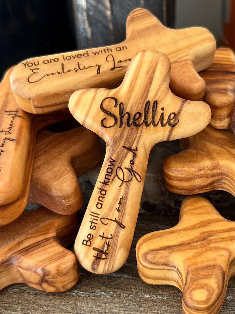 May include: A collection of wooden crosses with rounded edges and a natural wood grain. One cross is engraved with the name "Shellie" and the phrase "Be still and know that I am God." Another reads "You are loved with an Everlasting Love."