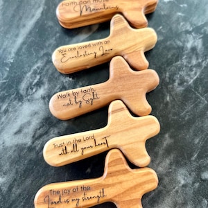 May include: Five wooden cross-shaped pocket prayer tokens with inspirational quotes engraved in black. The quotes include "Faith can move Mountains," and "Trust in the Lord with all your heart." The tokens are made of light-colored wood.