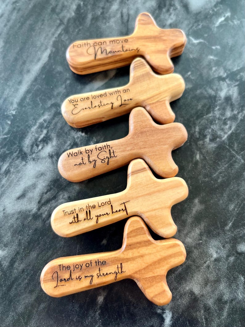 May include: Five wooden cross-shaped pocket prayer tokens with inspirational quotes engraved in black. The quotes include "Faith can move Mountains," and "Trust in the Lord with all your heart." The tokens are made of light-colored wood.