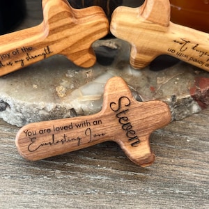 May include: Wooden crosses with rounded edges, featuring engraved text. One cross reads "You are loved with an Everlasting Love" and "Steven." Another says "joy of the Lord is my strength." The crosses are made of light brown wood.