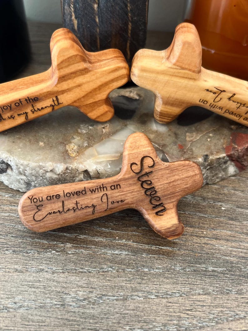 May include: Wooden crosses with rounded edges, featuring engraved text. One cross reads "You are loved with an Everlasting Love" and "Steven." Another says "joy of the Lord is my strength." The crosses are made of light brown wood.