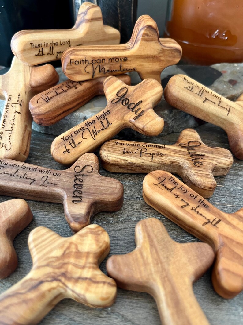 May include: A collection of wooden crosses with various engraved inspirational phrases and names. The crosses are made of light and dark brown wood, with visible grain patterns. Some phrases include "Trust in the Lord," and "Faith can move mountains."