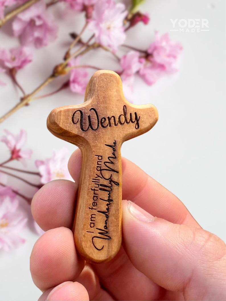 May include: A wooden cross with the name "Wendy" engraved on the top and the phrase "I am fearfully and wonderfully made" engraved on the bottom. The cross is held in a hand, with pink flowers in the background.