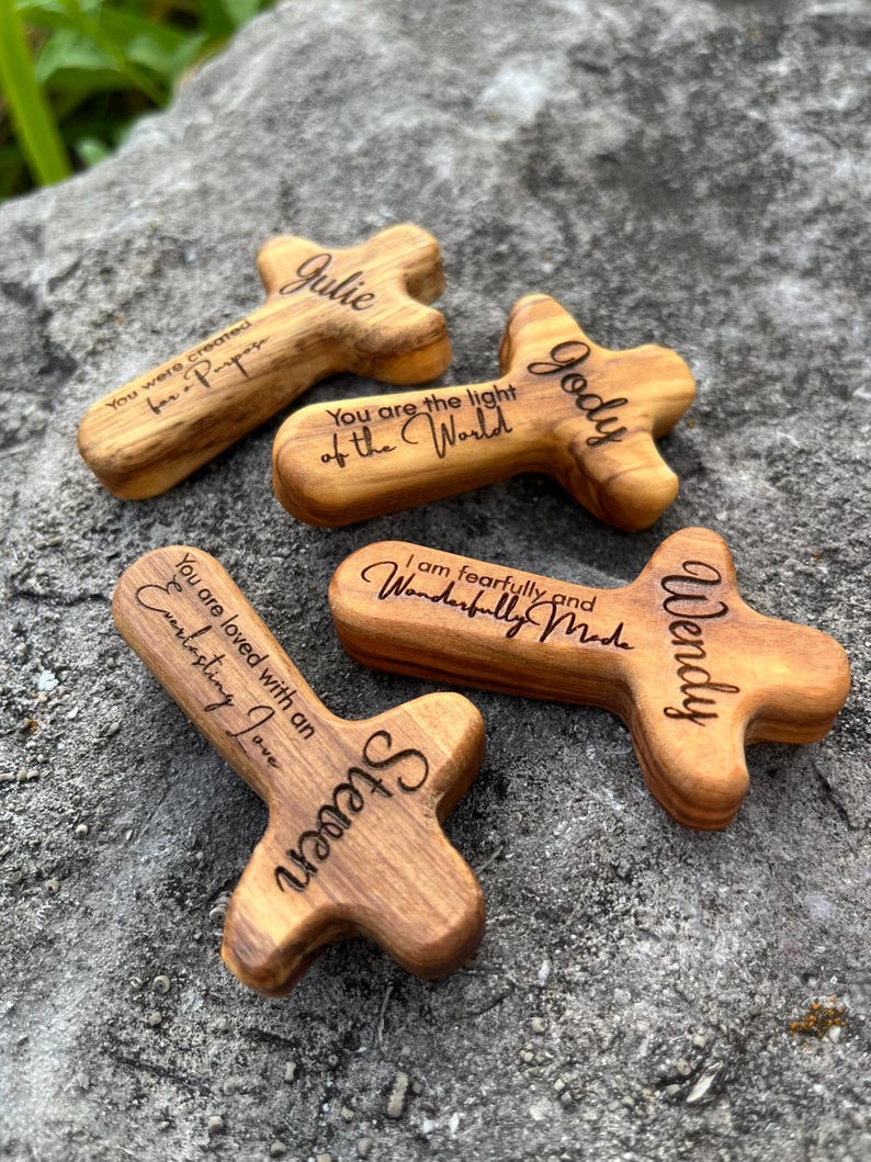 May include: Four wooden cross-shaped keepsakes with engraved names and inspirational quotes. The crosses are made of light brown wood and rest on a gray stone surface. The names Julie, Jody, Wendy, and Steven are visible.
