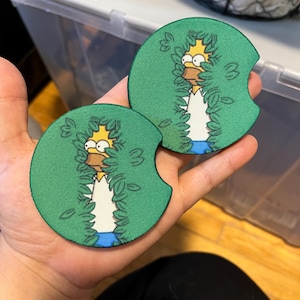 May include: Two round green drink coasters featuring a cartoon image of Homer Simpson peeking through green leaves. Homer is wearing a white shirt and blue trousers. The coasters have a small notch on one side.