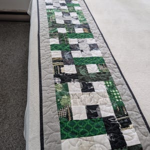 May include: A long, rectangular quilted bed runner featuring a patchwork design of green, white, black, and grey fabric squares. The runner has a central section with varied patterns and solid colours, bordered by a light grey quilted fabric edge.