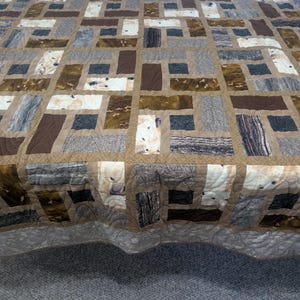 May include: A patchwork quilt featuring a grid of rectangular fabric pieces in shades of brown, tan, grey, and cream. The design incorporates various textures and patterns, including solid colours, mottled prints, and wood grain motifs, all joined by tan sashing.
