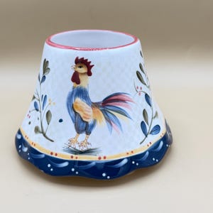 May include: A white ceramic lamp shade with a flared, scalloped base featuring a hand-painted rooster in shades of blue, red, and yellow. The design includes green leafy sprigs and a dark blue border with white dots, finished with a thin red rim.