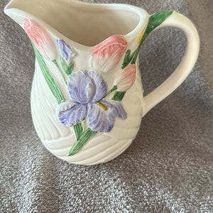 May include: White ceramic creamer with a floral design. The design features pink tulips and a purple iris with green leaves.