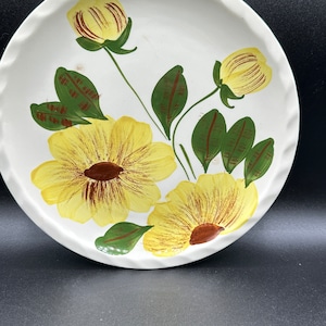 May include: A white ceramic plate with a yellow and green floral design. The flowers are painted in a stylized manner with brown accents.