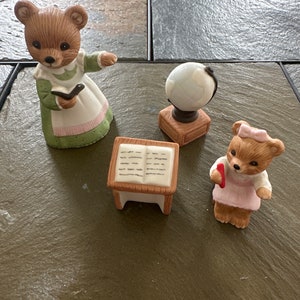 May include: A set of three ceramic bear figurines depicting a teacher, a student, and a globe. The teacher bear is wearing a green dress with a white apron and holding a book. The student bear is wearing a pink dress and holding a red heart. The globe is on a wooden stand.