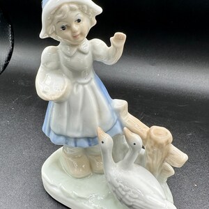 May include: A porcelain figurine of a girl in a blue and white dress and hat, feeding two white geese near a wooden fence.