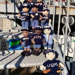 May include: A group of crocheted USPS mail carrier figurines, some wearing blue and white uniforms, others wearing blue and brown uniforms, standing on a set of stairs. One figurine is riding a small black bicycle.
