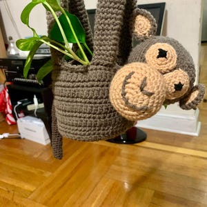 Puede incluir: Un macetero de mono de crochet marrón colgado de una varilla de madera. El mono tiene una cara beige y ojos negros. Una planta verde está creciendo dentro del macetero.