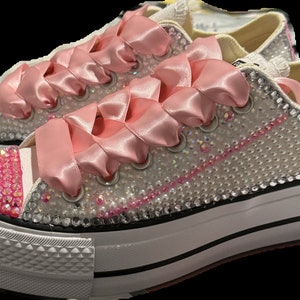 Puede incluir: Zapatillas blancas de plataforma con cordones de satén rosa y un diseño brillante rosa y blanco. Los zapatos están cubiertos de diamantes de imitación.