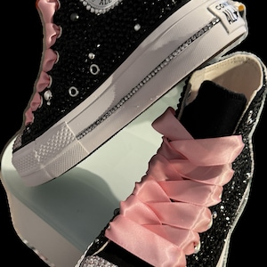 May include: Black high-top Converse sneakers with pink satin laces and a full-coverage crystal embellishment. The Converse logo is visible on the side of the shoe.