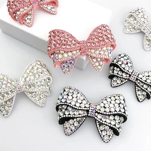 May include: Five rhinestone bow hair clips in various colors, including pink, white, and black. The bows are all made of rhinestones and have a shiny finish.