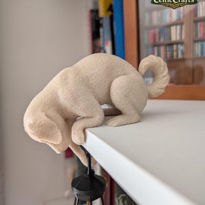 Può includere: Una statuetta di cane color crema con una superficie testurizzata, raffigurata in posizione accovacciata sul bordo di uno scaffale. Il cane tiene una piccola lanterna nera con una luce gialla brillante sospesa dalle zampe anteriori e ha una coda arricciata.