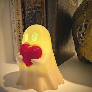 May include: A small, off-white, ghost-shaped figurine with glowing eyes, holding a solid red heart in its hands. The figurine has a smooth, matte texture and is positioned upright, emitting a soft, warm light from within its hollow structure.
