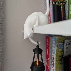 May include: A white cat figurine perched on the edge of a shelf, with its tail hanging down to hold a black lantern. The lantern features a small, glowing orange flame-shaped light inside its frame. The cat is positioned above a row of books.