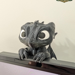 May include: A black 3D printed dragon figure with large white eyes and textured skin, designed to perch on the top edge of a computer monitor. The creature has small wings, sharp claws gripping the screen frame, and a smooth, matte finish.