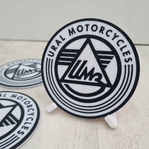May include: A white round coaster with a black and white logo that says "URAL MOTORCYCLES" in a circle around a triangle with the letters "UM" inside.