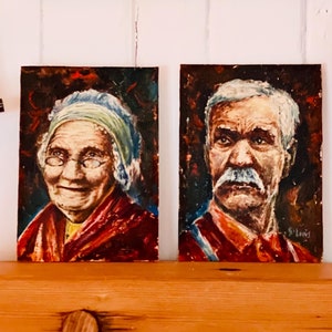 May include: Two framed oil paintings of older people. The painting on the left shows a woman with a white headscarf and glasses smiling. The painting on the right shows a man with a white beard and mustache looking straight ahead. Both paintings have a dark background.