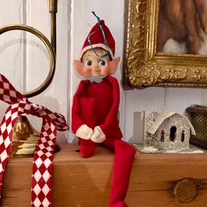 May include: A red elf doll with a pointed hat and white cuffs sits on a wooden shelf. The elf has a smiling face with blue eyes and large ears. A red and white chequered ribbon is tied to a brass horn.