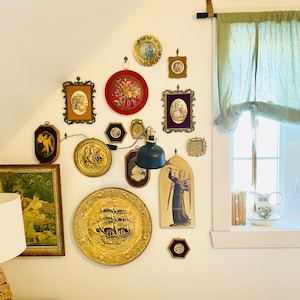 May include: A white wall with a collection of framed artwork and decorative plates. The artwork includes a variety of subjects, including religious figures, landscapes, and still lifes. The plates are made of gold metal and feature nautical scenes. A black lamp with a white shade hangs from the ceiling.