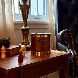 May include: A wooden tobacco jar with brass accents and the word "TOBACCO" on a label. The jar is cylindrical and sits on a wooden table with chess pieces and a book. A brass lamp and a leather armchair are in the background.