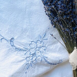 Könnte beinhalten: Ein weißes Leinentextil mit hellblauer Blumenstickerei in der Ecke, kombiniert mit einem Bündel getrockneten Lavendels. Die Stickerei zeigt ein zartes Blumen- und Blattdesign. Der Lavendel ist mit einem naturfarbenen Band gebunden.