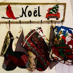 Peut inclure: Une pancarte en bois avec le mot "Noël" peint en noir. La pancarte a des crochets pour accrocher des chaussettes. Il y a cinq chaussettes accrochées aux crochets, chacune avec un motif différent. Une chaussette a un motif de Père Noël.