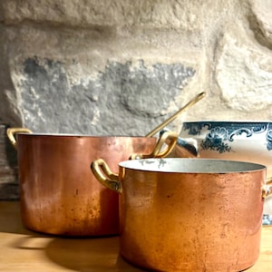 May include: Two copper cooking pots with brass handles are displayed on a wooden surface. The pots have a polished, reflective finish and vary in size. A blue and white patterned ceramic bowl is partially visible in the background, suggesting a kitchen or dining setting.