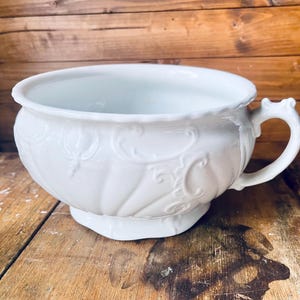 Könnte beinhalten: Ein weißer Keramik-Pflanztopf mit Griff und geprägten Verzierungen. Der Pflanztopf hat eine breite, offene Schalenform und steht auf einem kleinen, achteckigen Sockel. Der Griff ist gebogen und hat ein dekoratives Design.