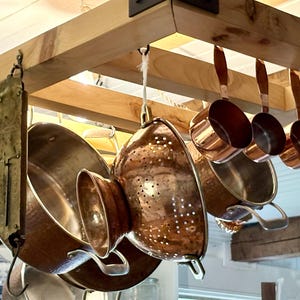 May include: A collection of copper kitchenware hanging from a wooden rack, including a large hammered copper colander, a deep copper pot with two side handles, and several small copper measuring cups with long handles, all suspended by metal hooks.