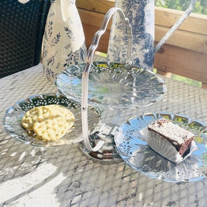 May include: A three-tiered, silver serving tray with a handle, holding crackers and a piece of cake. The tray has a decorative, scalloped edge. The background includes a white tablecloth and decorative vases.