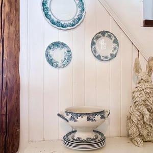 May include: A collection of vintage white and teal ceramic tableware. Includes decorative plates, a soup tureen, and a stack of plates. The plates are mounted on a white wall, and the tureen sits on a white cabinet. A decorative rabbit statue is also present.