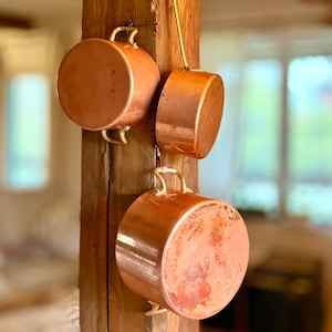 May include: Three copper pans with brass handles suspended from a wooden beam. The pans vary in size and form, with a bright, reflective surface. The copper has a warm, reddish-brown tone, and the pans appear to be vintage or antique, adding a rustic charm.