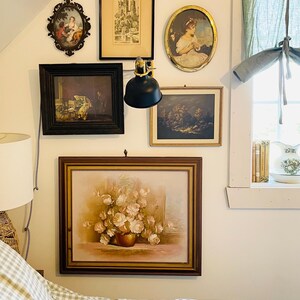 May include: A bedroom with a white wall and a bed with a grey and white checked duvet. The wall is decorated with framed artwork, including a floral painting, a landscape painting, and a portrait. There is a black lamp on the wall.