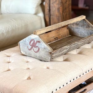 May include: A weathered wooden toolbox with a gray and white finish, featuring the number "95" in red on the side. The toolbox is resting on a tufted ottoman. The background includes a white sofa and wooden beams.