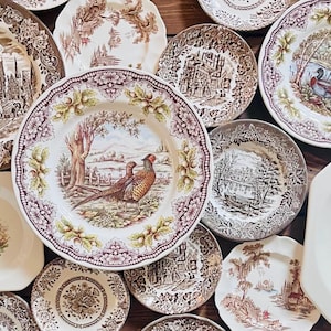 May include: A collection of vintage ceramic plates with brown and white floral patterns. The plates feature various scenes, including a pheasant, a house, and a boat.