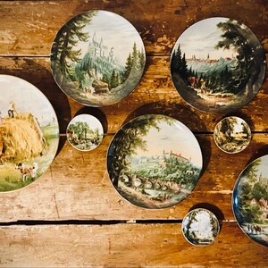 May include: A collection of eight decorative plates featuring various painted scenes. The plates are round and have a white background with green, brown, and blue accents. The scenes include a hay wagon, a castle, a city, and a forest.