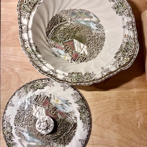 Könnte beinhalten: Eine weiße Keramikschüssel und ein Deckel mit Landschaftsdesign. Die Schüssel hat einen gewellten Rand mit einem braunen und grünen Blumenrand. Der Deckel hat ein passendes Design und einen weißen Knopf mit floralem Muster.