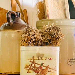 May include: A selection of vintage kitchenware, including a ceramic jug, a stoneware crock, and a decorative tin. The tin displays a winter scene with pheasants and a wooden fence. Dried flowers are placed in front of the tin.