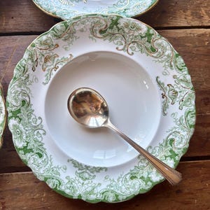 May include: A white soup bowl with a green and gold floral design and a silver spoon. The bowl has a scalloped edge. The spoon is resting inside the bowl. The bowl is on a wooden surface.