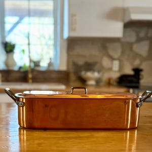 May include: A copper rectangular roasting pan with a lid and two side handles. The pan is on a wooden surface. The background shows a kitchen with a window and a stone backsplash.