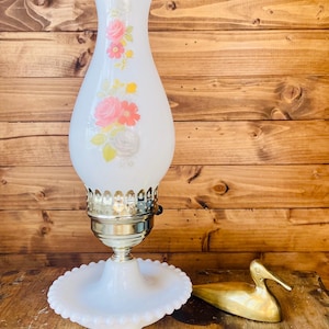 May include: A vintage white glass lamp with a floral design featuring pink and red roses. The lamp has a brass base and a scalloped edge. A brass duck figurine sits beside the lamp on a wooden surface.