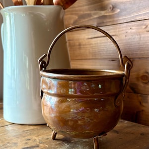 May include: A small, antique copper cauldron with a handle and three small legs. The cauldron has a hammered texture and a rich, warm brown colour. A white ceramic jug and wooden kitchen utensils are in the background.